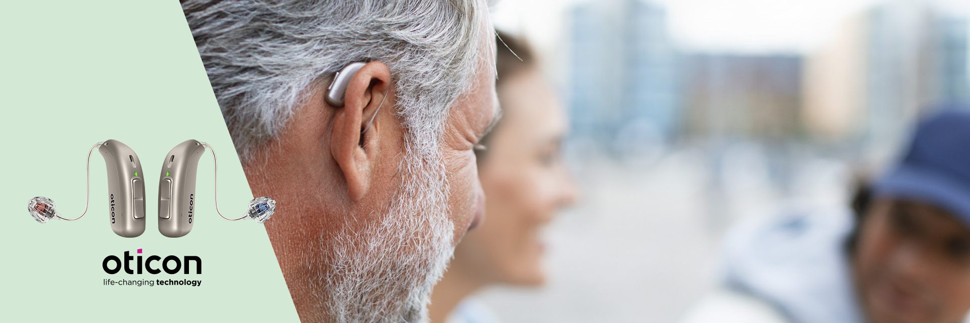 A silver-haired man wears an Oticon hearing aid discreetly behind his ear, with blurred people and urban architecture in the background. Text reads: 'Oticon - Life-changing technology.' Two Oticon hearing aids are displayed on a mint-green background.