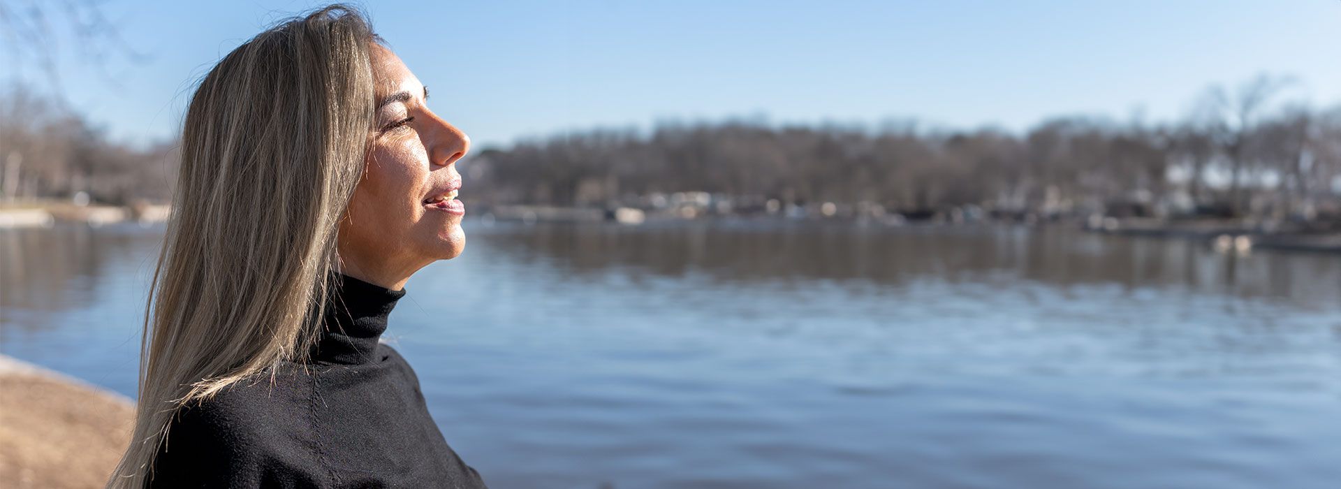 Eine Frau mit langen, blonden Haaren trägt einen schwarzen Rollkragenpullover, blickt auf einen ruhigen See an einem klaren, sonnigen Tag. Im Hintergrund sind kahle Bäume.