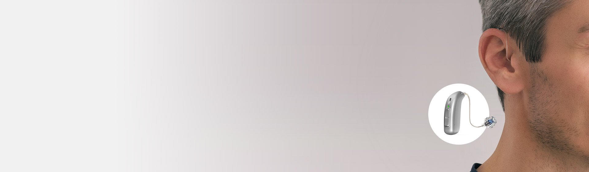 Un uomo adulto con capelli grigi mostra il profilo destro del viso; un apparecchio acustico retroauricolare grigio (modello Audika) è evidenziato su sfondo bianco sfumato a sinistra.