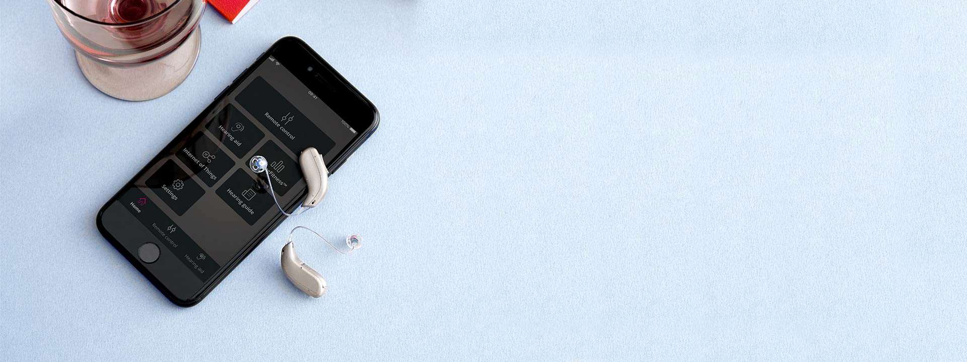 Due apparecchi acustici moderni sono posati su uno smartphone con schermo acceso che mostra un’app per il controllo degli apparecchi. Sullo sfondo, un tavolo chiaro con un bicchiere.