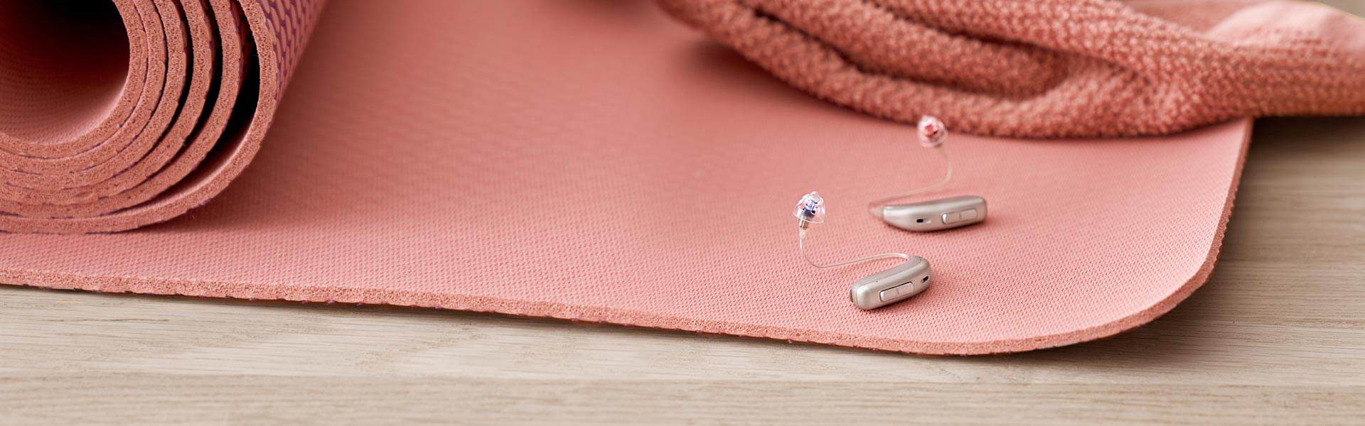 Zwei moderne Hörgeräte auf einer zusammengerollten, rosafarbenen Yogamatte.