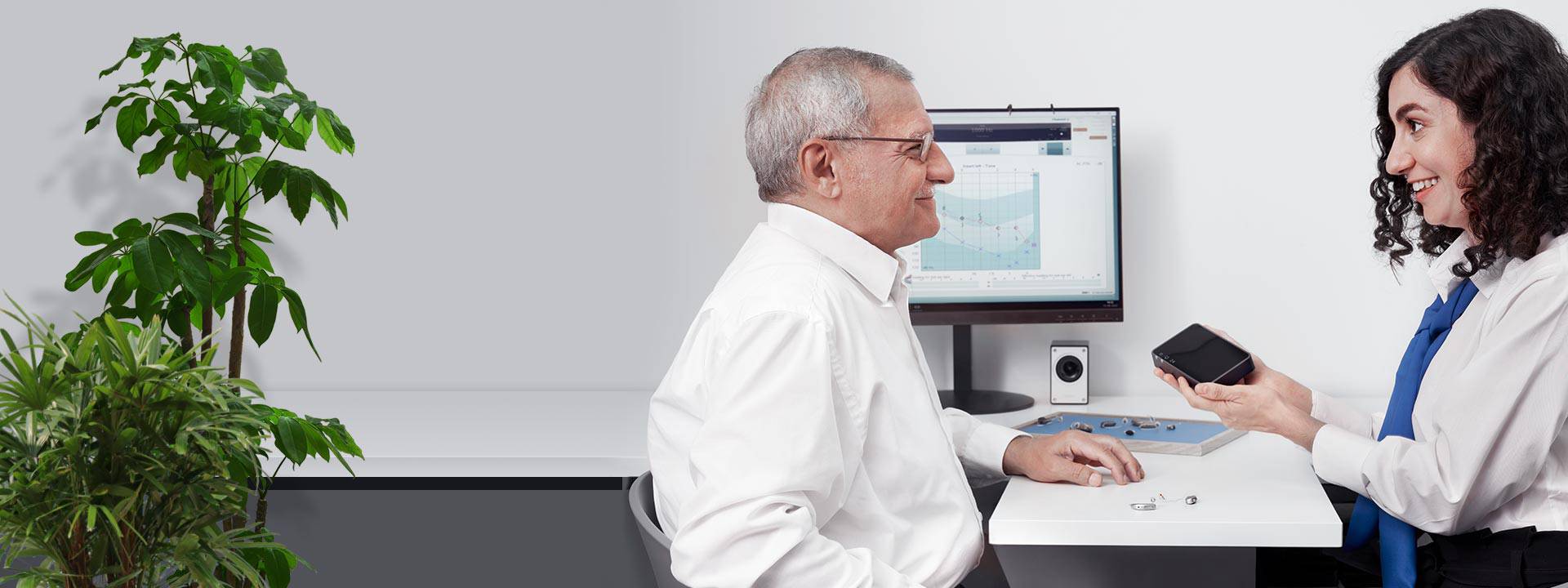 Man consulting with an audiologist while reviewing hearing aid accessories and data on a computer screen.