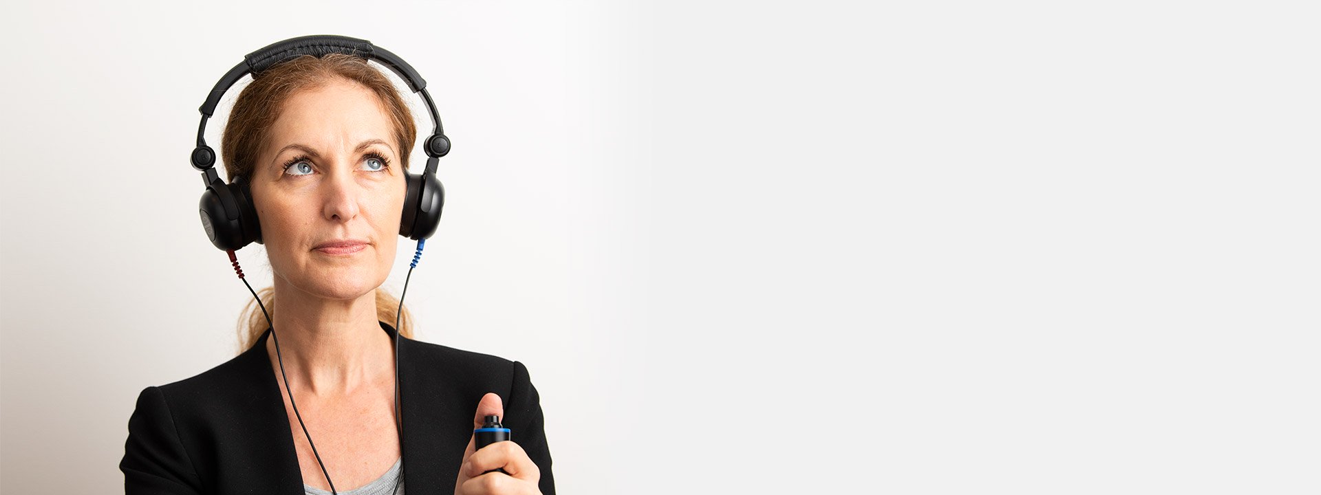 [GEN] A person wearing over-ear headphones holds a handheld button device, likely for a hearing test, against a plain white background, suggesting a clinical hearing assessment setting. No visible text or brand logos.