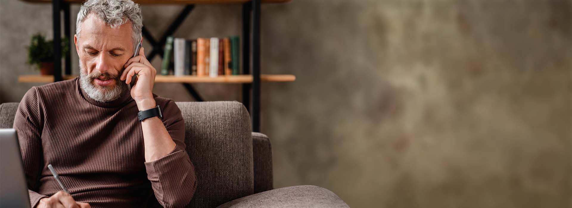 Man with gray hair talking on the phone while sitting on a sofa, with a bookshelf in the background.