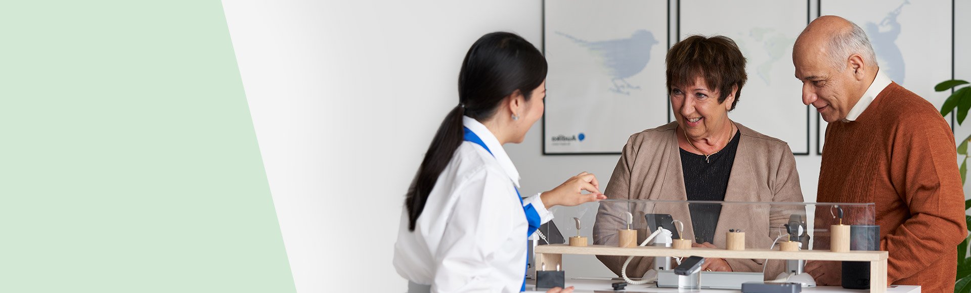 Especialista em audiologia orienta um casal num centro auditivo Audika. Sobre a mesa, há aparelhos auditivos em exposição, com foco no cuidado auditivo e saúde auditiva. Logo Audika visível.