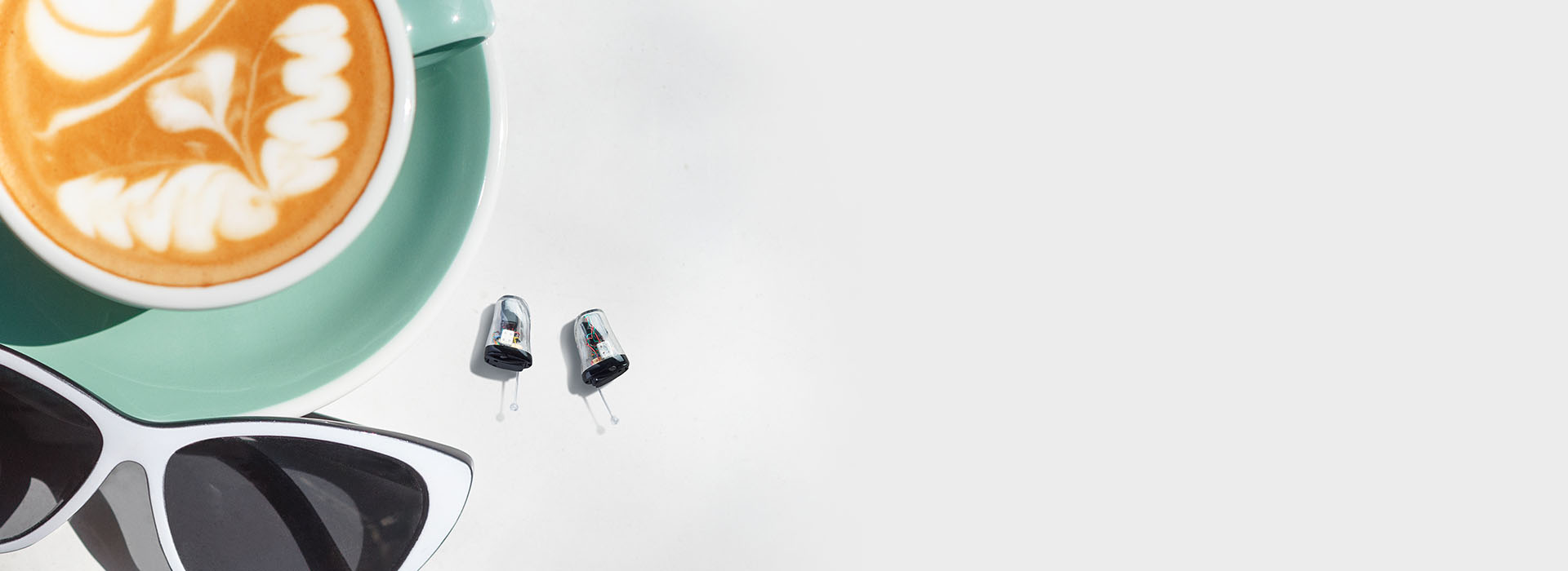[GEN] Two sleek, silver hearing aids sit on a white surface next to black sunglasses and a green cup holding a latte with intricate foam art, creating a stylish, modern setting.