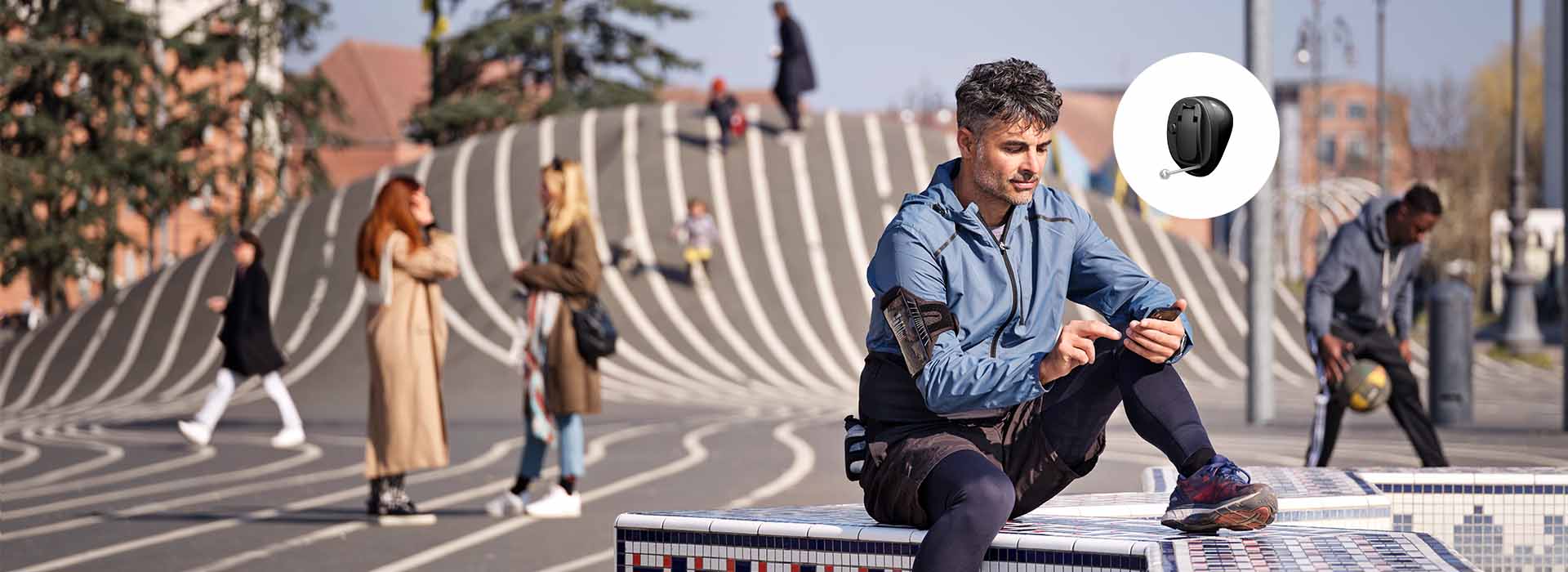Homem sentado em um banco mosaico, utilizando smartphone. Cenário urbano com pessoas interagindo em praça moderna de design listrado. Círculo destaca um aparelho auditivo discreto, modelo intracanal.