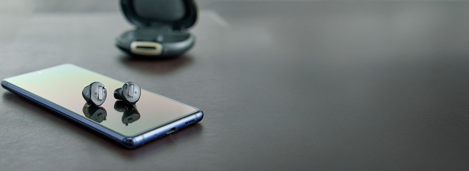 To høreapparater ligger på en smartphone med skærmen opad. I baggrunden ses et åbent opladningsetui. Scenen er sat på en grå bordflade i minimalistisk stil.