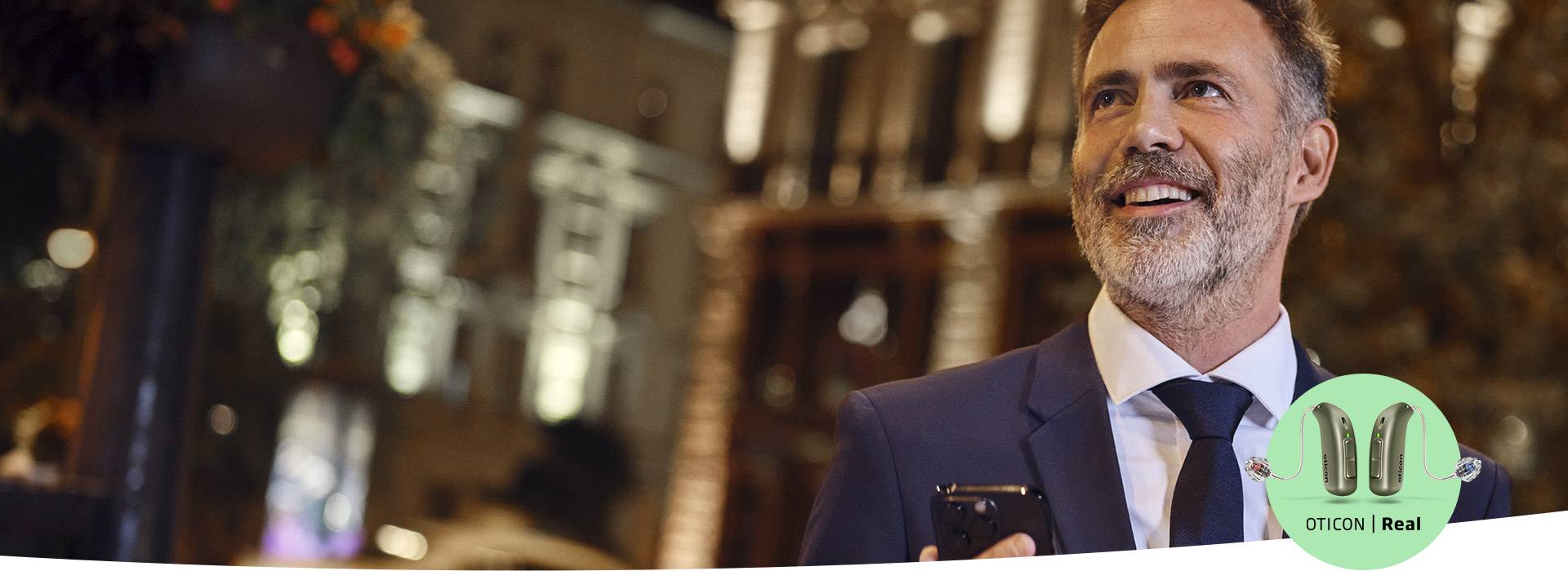 Mężczyzna w garniturze trzyma telefon, znajduje się w oświetlonej miejskiej scenerii nocą. W prawym dolnym rogu widoczne logo Oticon Real z wizerunkiem aparatów słuchowych.