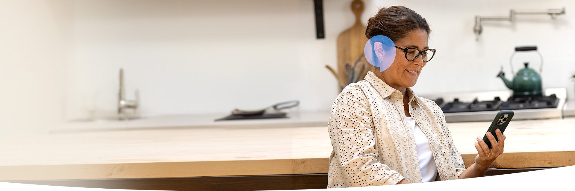 Une femme portant un appareil auditif examine un smartphone dans une cuisine lumineuse avec un plan de travail en bois, une bouilloire verte sur la cuisinière et un évier en arrière-plan.