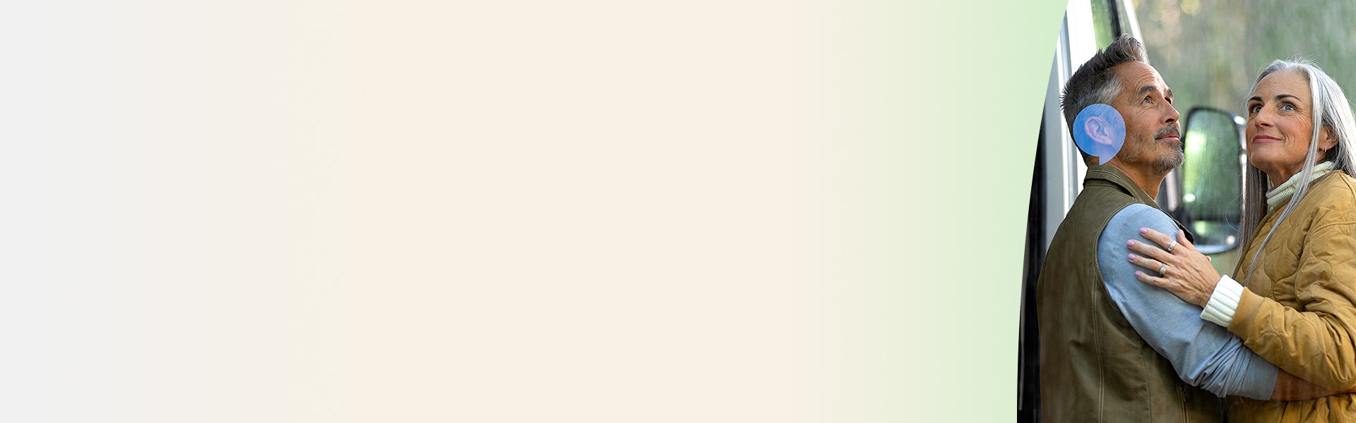 En mand med et høreapparat krammer en kvinde tæt ved en hvid bil i en udendørs, skovrig kontekst. Manden ser ud til at bruge høreteknologi fra Audika.