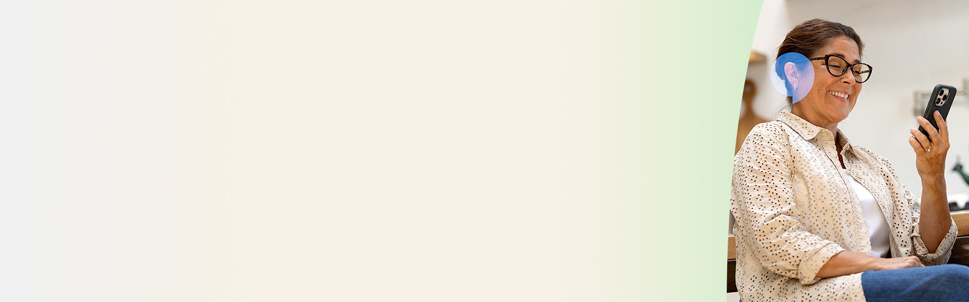 Une femme portant des lunettes et un appareil auditif regarde son smartphone, assise dans un environnement lumineux.