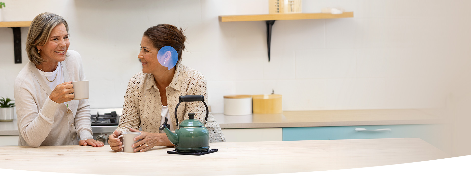 Deux femmes discutent en buvant du café dans une cuisine moderne lumineuse; une d'elles porte un appareil auditif discret. Une théière verte repose sur le plan de travail.