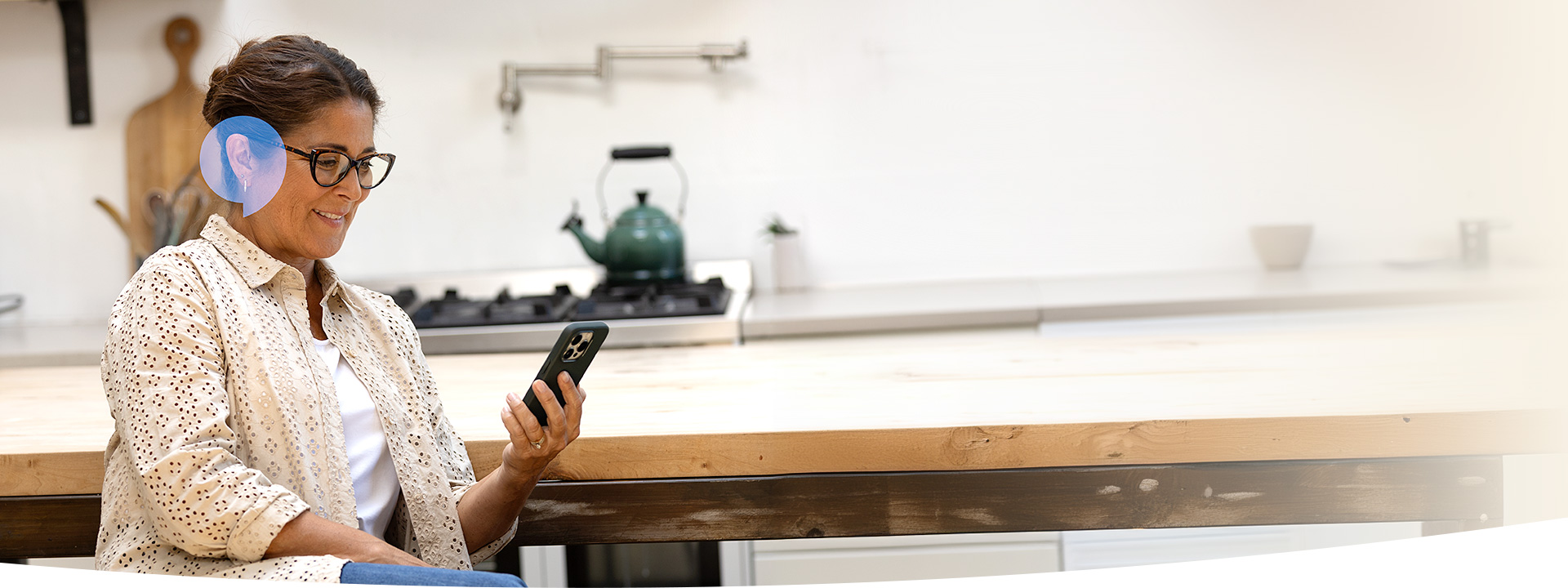 Une femme portant un appareil auditif consulte son smartphone dans une cuisine moderne, équipée d’une bouilloire verte sur une cuisinière. Elle est assise près d’une table en bois clair.