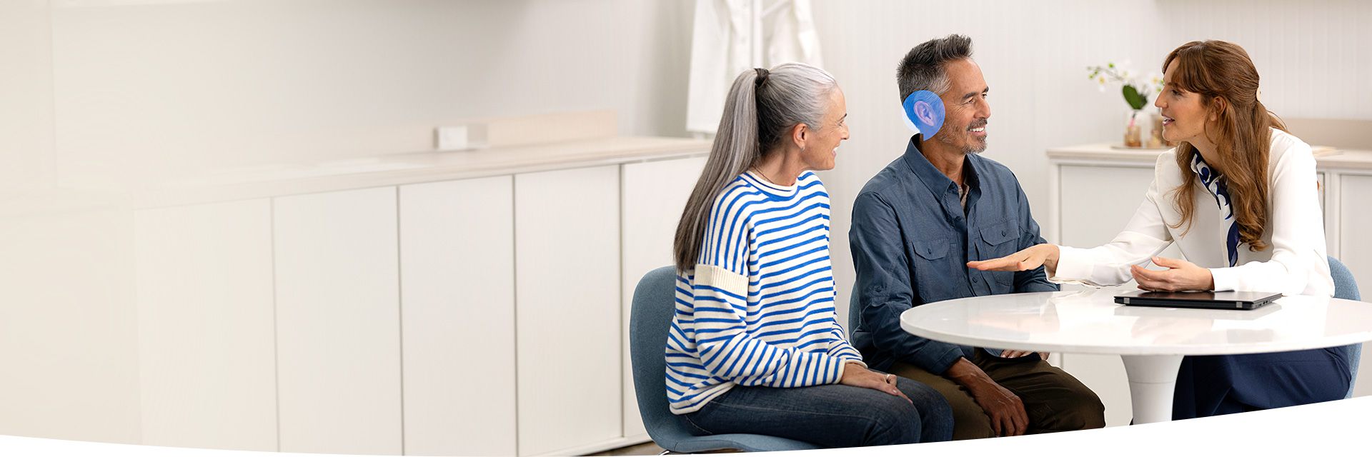 Een audioloog spreekt met een man, die een hoortoestel draagt, en een vrouw aan een ronde tafel in een moderne, lichte hoorkliniek met witte kasten.
