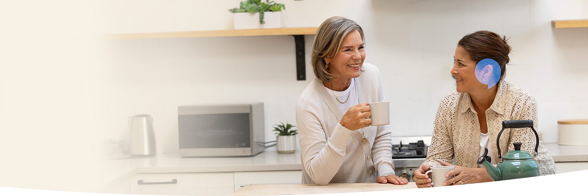 Two women enjoying tea in a kitchen, one wearing a hearing aid highlighted with a blue overlay.