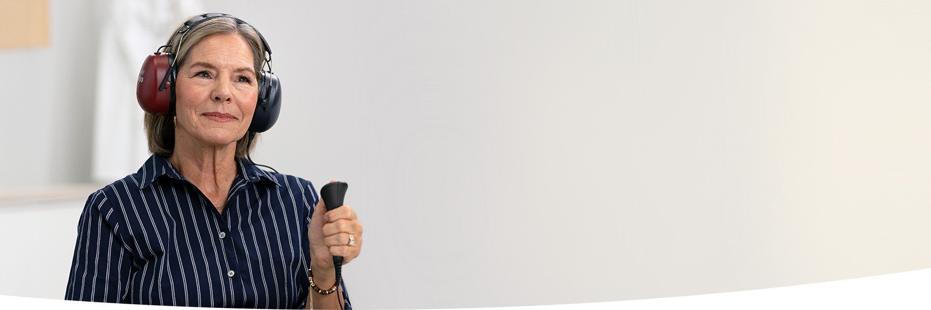 Uma mulher com fones de ouvido protetores realiza um teste auditivo em uma clínica, segurando um botão de resposta manual. Fundo claro e ambiente típico de avaliação auditiva.