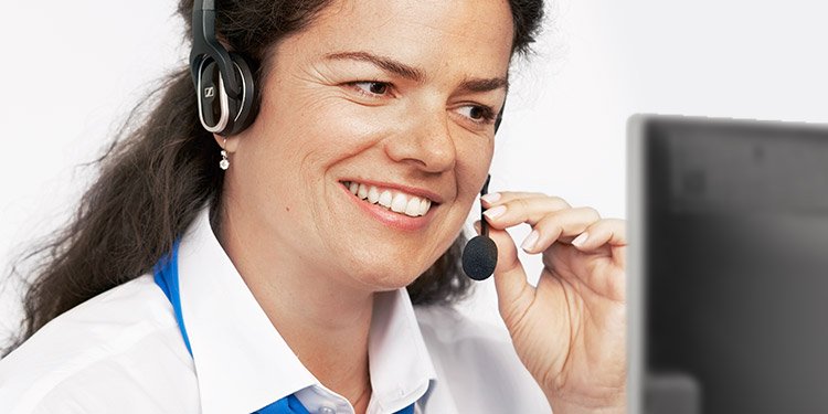 Uma profissional de audiologia, usando um headset preto da marca Sennheiser, interage em frente a um monitor em um ambiente clínico. O foco sugere suporte num centro auditivo.