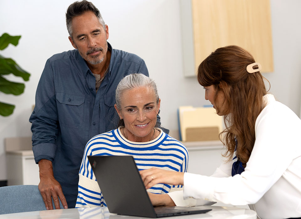 Une personne âgée assise, mains croisées, échange avec une femme aux cheveux longs dans un environnement lumineux d'intérieur, possiblement un cabinet spécialisé en tests auditifs ou conseil sur les appareils auditifs.