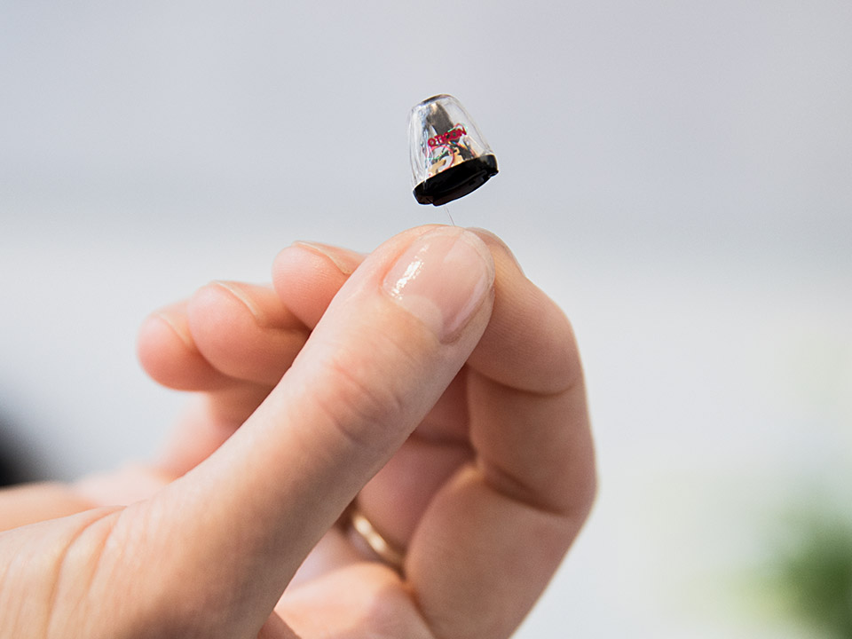 A discreet in-the-ear hearing aid held gently between fingertips, featuring Oticon branding. The device is showcased in clean, well-lit surroundings, emphasizing advanced hearing care and precision technology.