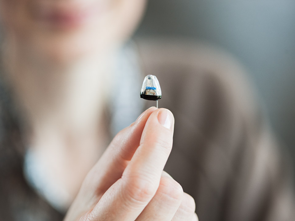 Osoba trzyma w palcach mały, dyskretny aparat słuchowy marki Oticon. Urządzenie znajduje się na pierwszym planie, w tle rozmazane kontury twarzy i neutralne otoczenie.