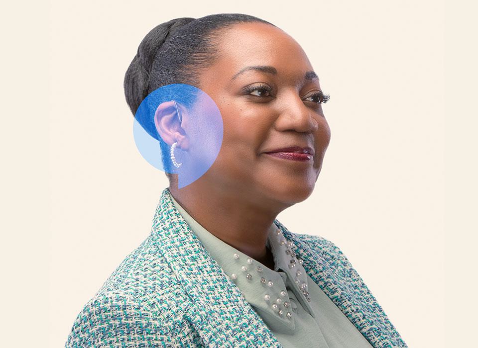 A person with a blurred face wears a discreet behind-the-ear hearing aid, highlighted by a blue accent, in a professional setting. They are dressed in a green tweed jacket and pearl-embellished shirt, suggesting a focus on hearing care services.