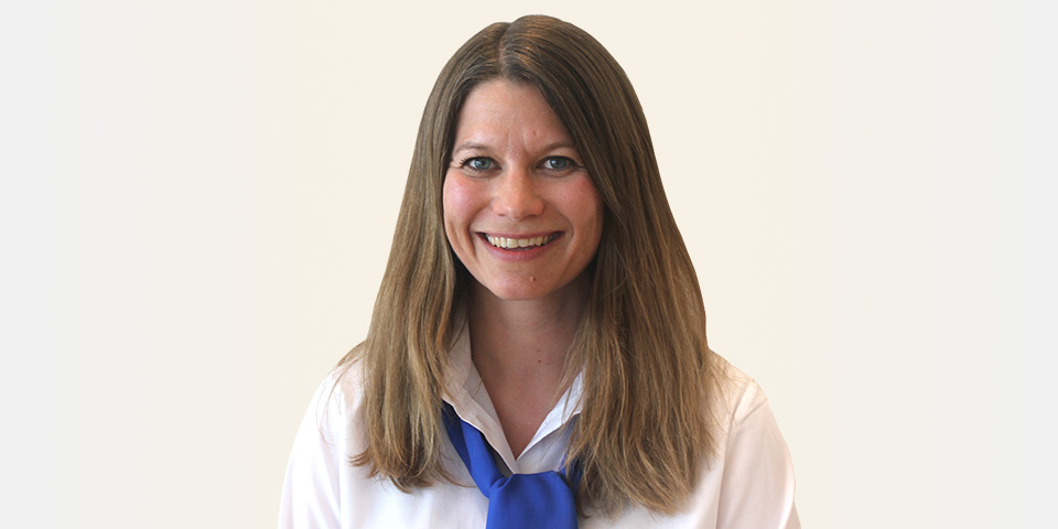 Une personne en chemise blanche avec une cravate bleue se tient devant un fond uni. Le visage est flouté, mettant en avant l’uniforme professionnel, peut-être dans un contexte de clinique auditive.