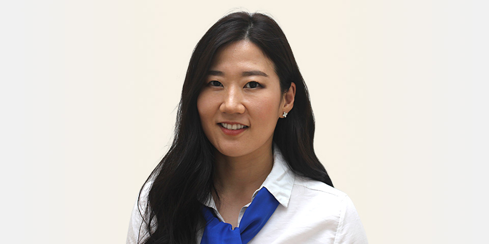 Une femme en chemise blanche avec un foulard bleu, cheveux longs et noirs, porte des boucles d’oreilles. L’arrière-plan est uni et clair. Aucun appareil auditif discernable.