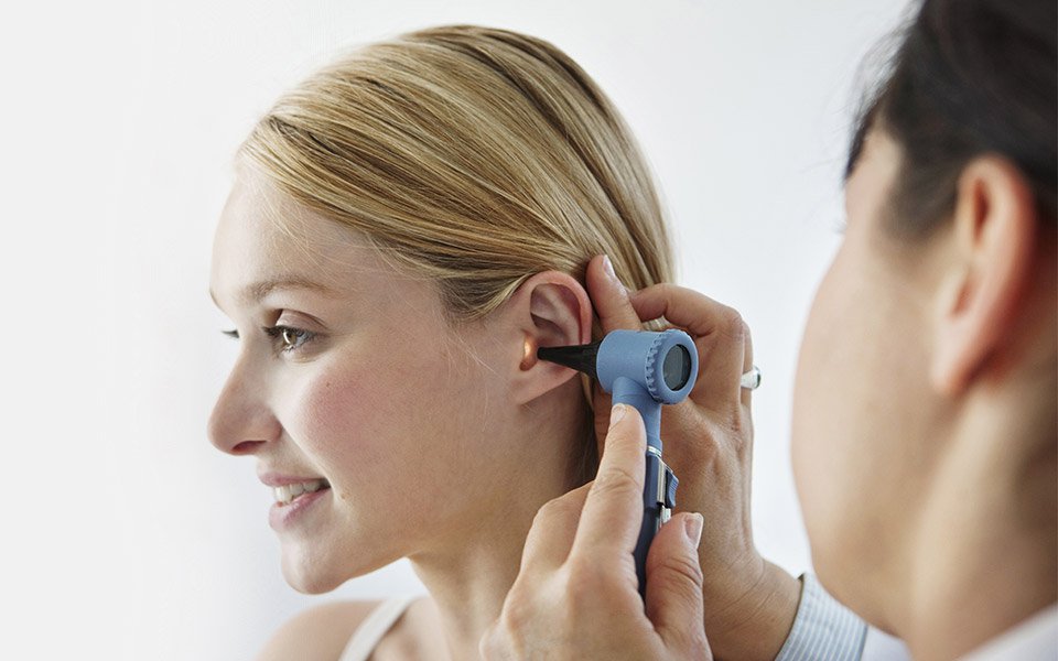 An audiologist conducts an ear examination using an otoscope on a blonde-haired individual in a clinical setting, emphasizing professional hearing care services like hearing tests and assessments for potential hearing aid needs.