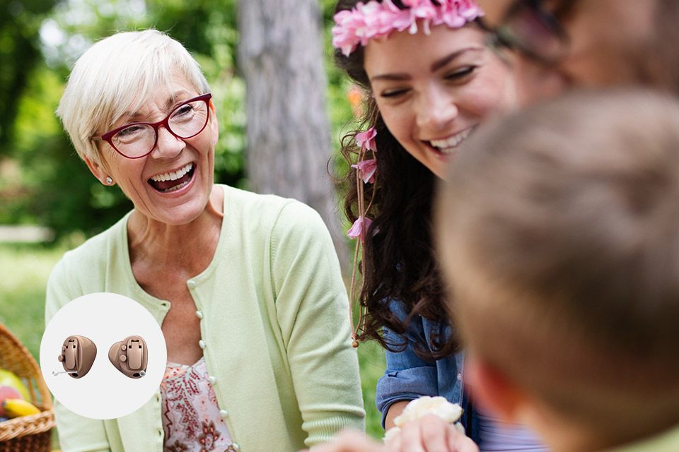 Ældre kvinde iført pastelgrøn cardigan smiler og interagerer i en have med familie. Indsat billede viser to beige høreapparater. Fokus på hørelse og familieliv i naturlige omgivelser.