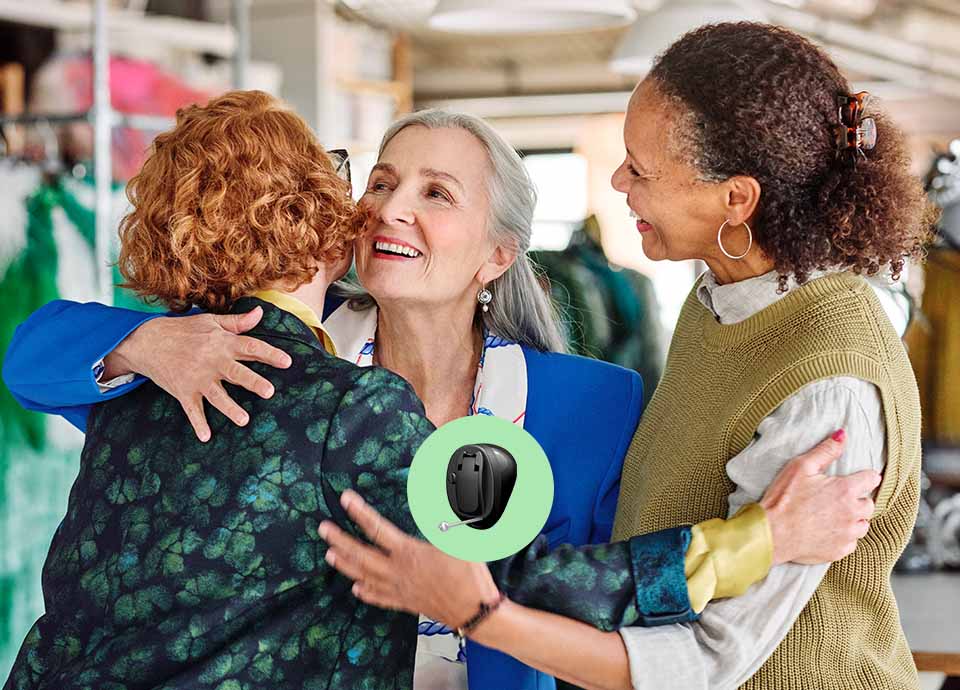 Three women embrace warmly in a brightly lit indoor setting, surrounded by colorful garments. A prominently displayed Oticon hearing aid is featured within a green circle overlay, highlighting advanced hearing care solutions.