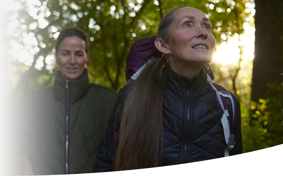 To personer iført udendørsjakker og rygsække går i en skov med sollys, der trænger gennem træerne. Ingen synlig tekst, høreapparater eller andre Audika-relaterede elementer.