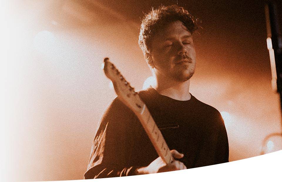 A guitar player performs on stage, holding an electric guitar under warm, ambient lighting, creating a hazy, intimate concert atmosphere.