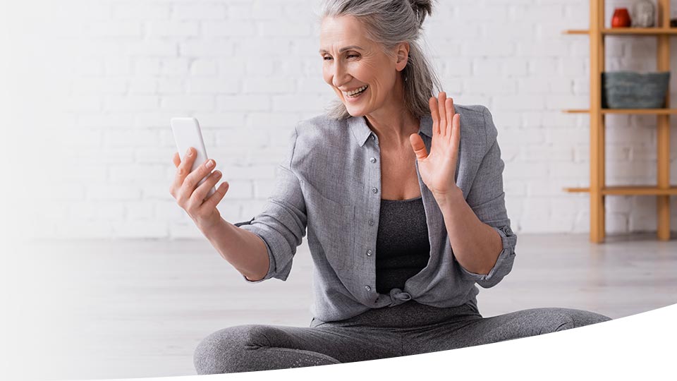 A seated woman with gray hair uses a smartphone, raising her hand as if greeting someone during a video call. The bright room has a white brick wall and wooden shelving.