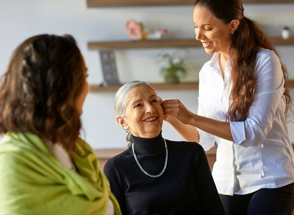 Uma profissional de saúde auditiva ajusta um aparelho auditivo em uma mulher de cabelo grisalho, enquanto outra mulher observa. O ambiente é iluminado, com estantes e plantas ao fundo.