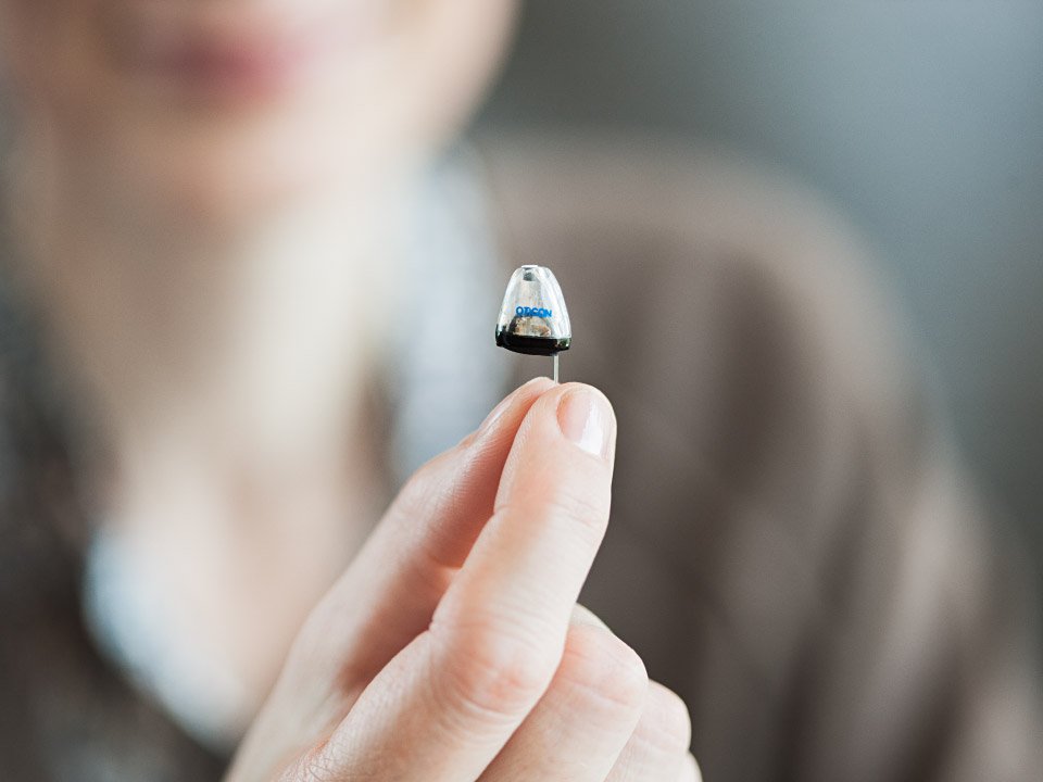 Osoba trzyma w palcach mały, dyskretny aparat słuchowy marki Oticon. Urządzenie znajduje się na pierwszym planie, w tle rozmazane kontury twarzy i neutralne otoczenie.