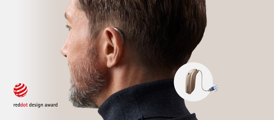 Um aparelho auditivo discreto, possivelmente da Oticon, é usado atrás da orelha de um homem em close, fundo neutro. Logotipo ’Red Dot Design Award’ aparece no canto esquerdo inferior.