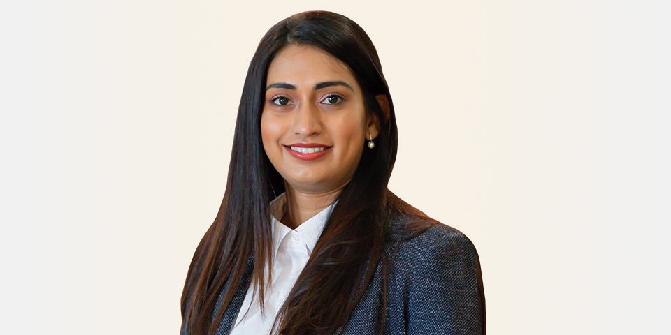 Une femme aux cheveux longs et bruns porte une veste formelle bleue et une chemise blanche, debout sur un fond uni. Le visage de la femme est flouté. Aucun texte ni appareil auditif identifiable n’est visible.