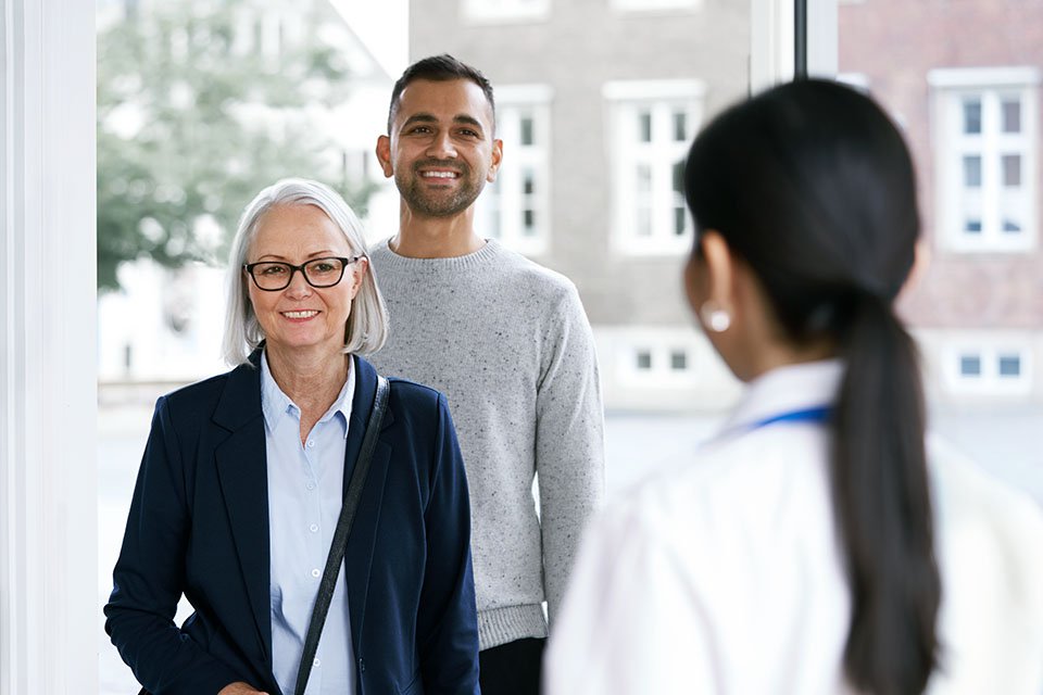 Audiologist greeting an older woman and a younger man at a hearing clinic.