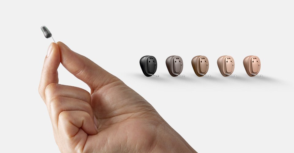 Uma mão segura um discreto aparelho auditivo intra-auricular. Ao lado, cinco aparelhos auditivos Oticon de diferentes cores estão alinhados sobre um fundo branco, representando opções disponíveis em clínicas auditivas.