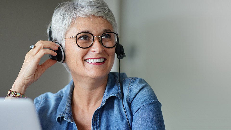 Starsza osoba z siwymi włosami nosi słuchawki z mikrofonem, trzymając je jedną ręką. Ubrana jest w dżinsową koszulę, siedząc przy biurku z laptopem w jasnym, prostym otoczeniu.