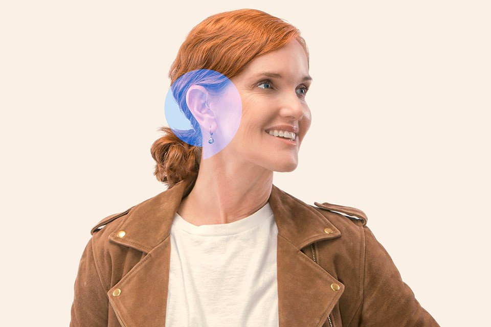 Una persona con cabello pelirrojo lleva un audífono detrás de la oreja izquierda. Viste chaqueta marrón y camiseta blanca, en un fondo beige neutro.