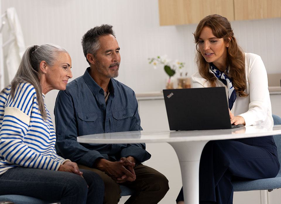 A hearing care specialist consults with an older couple at a round white table in a modern hearing clinic setting, using a Lenovo laptop. Orchids and neutral decor enhance the environment.