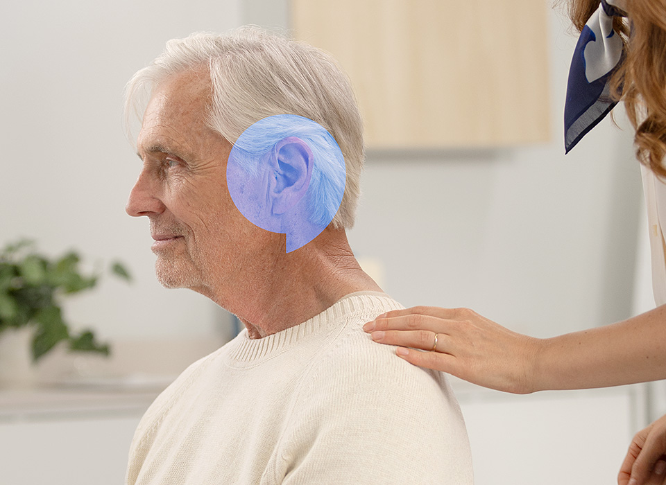 Un homme aux cheveux blancs, portant un pull beige, est assis dans un cadre de clinique auditive moderne. Une main féminine touche son épaule, suggérant soutien ou consultation. Aucune marque spécifique d’appareil auditif identifiable.