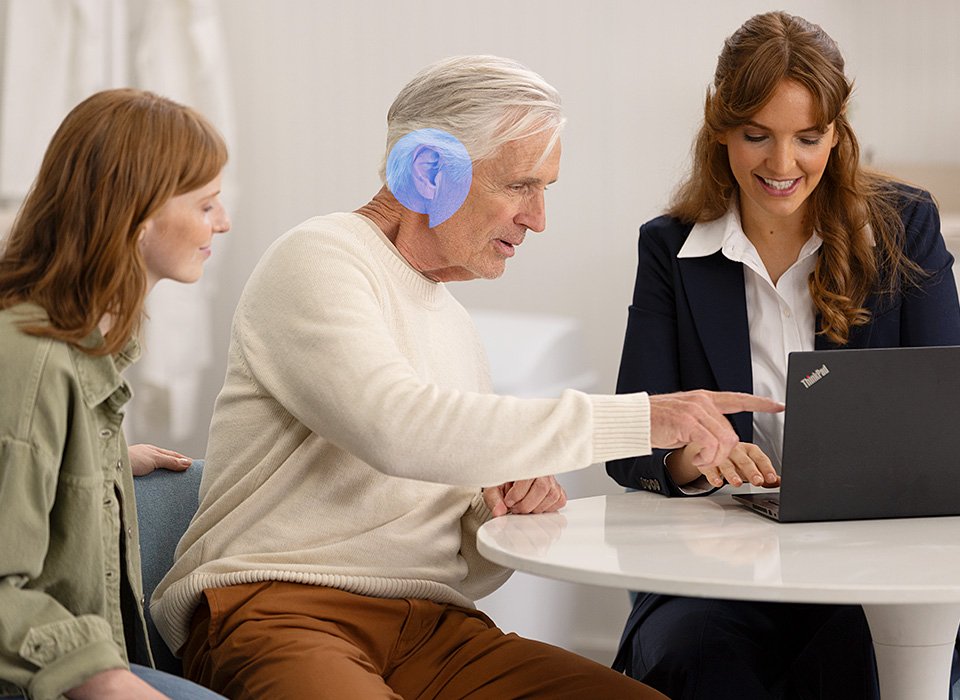 Un homme âgé portant un appareil auditif bleu interagit en pointant un écran d’ordinateur portable Lenovo ThinkPad, accompagné de deux femmes dans un environnement lumineux, possiblement une clinique auditive.