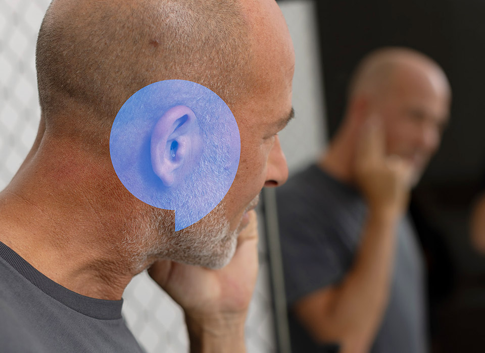 Un gros plan sur l’oreille d’un homme portant un appareil auditif discret. En arrière-plan flou, un autre homme se tient dans un environnement intérieur sobre. Aucun texte visible.