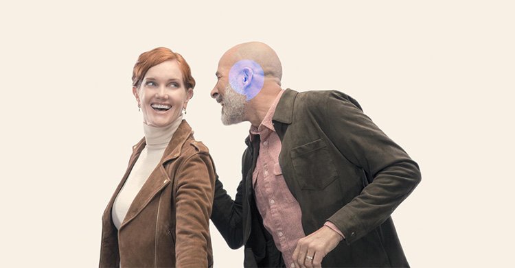 A man leans in closely while speaking to a woman, both dressed in brown and beige clothing against a plain beige background.