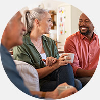 A group of individuals seated in a cozy living room holds mugs while conversing. The central figure, with gray hair, wears a green shirt, facilitating a casual, social environment. No text or identifiable hearing aid brands are visible.