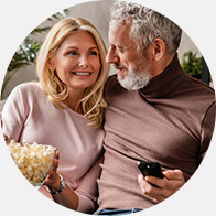 A couple sits closely on a sofa; the woman holds a bowl of popcorn while the man operates a remote control. The cozy setting suggests relaxation, possibly watching TV. No text visible.