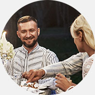 A man and woman are seated outdoors at a candlelit dinner table with floral arrangements, sharing a meal in an intimate and relaxed evening setting.